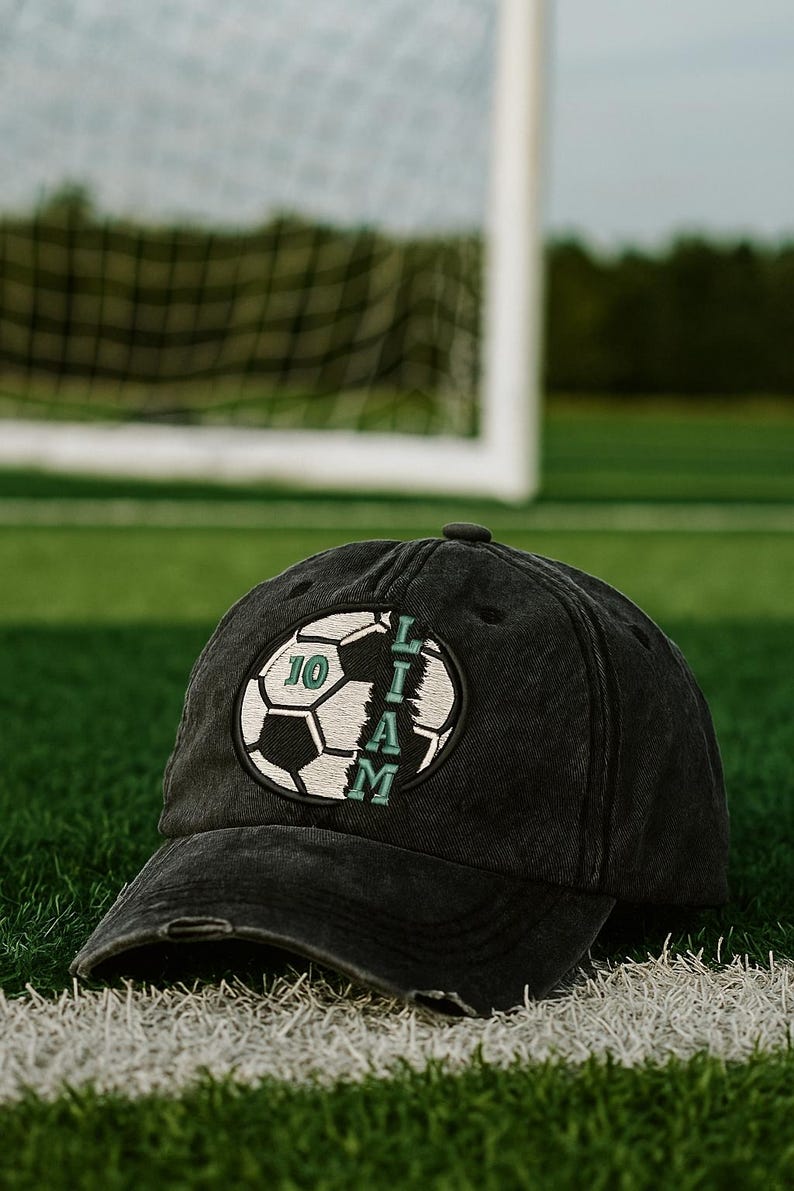 K&ouml;nnte beinhalten: Schwarze Baseballkappe mit Used-Look, mit einem Fu&szlig;ball-Design mit der Nummer 10 und dem Namen "LIAM" in Gr&uuml;n und Wei&szlig;. Die Kappe liegt auf einem gr&uuml;nen Feld mit einer wei&szlig;en Linie, mit einem Fu&szlig;balltor im Hintergrund.