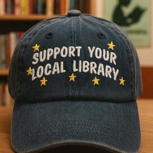 Peut inclure: Casquette de baseball bleu marine avec l'inscription brodée "SUPPORT YOUR LOCAL LIBRARY" en blanc, ornée d'étoiles jaunes. Le chapeau est en tissu doux et possède une visière incurvée. Un accessoire élégant pour les amateurs de livres.