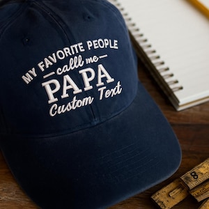 May include: Navy blue baseball cap with white embroidered text that reads "MY FAVORITE PEOPLE call me - PAPA Custom Text". The hat is on a wooden surface with a notepad and pencil.