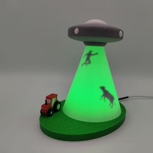 May include: A novelty lamp shaped like a grey UFO hovering over a green base. The lamp emits a bright green light in a cone shape, featuring dark silhouettes of a person and a cow being abducted. A small red and black tractor sits on the base.