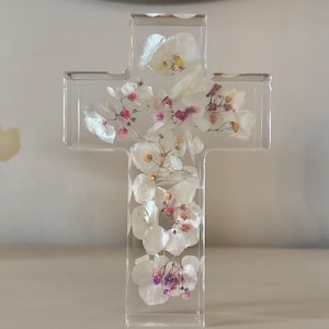 May include: A clear resin cross filled with white and pink dried flowers. The cross is standing upright on a light-colored surface. The flowers are arranged throughout the cross, creating a delicate and natural aesthetic.