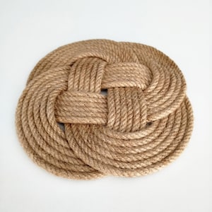 Può includere: Un tappetino rotondo a tema nautico realizzato con spessa corda color naturale. La corda è strettamente intrecciata in un motivo a nodo decorativo, creando una superficie strutturata. Il tappetino è circolare e poggia su uno sfondo bianco.