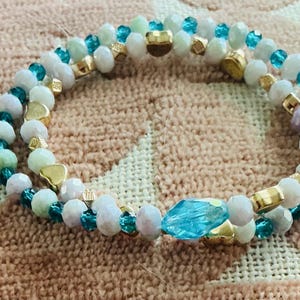 May include: A double-strand beaded bracelet featuring white, light green, and turquoise faceted beads interspersed with small gold-toned heart-shaped charms and a single larger, elongated, translucent blue crystal bead as a central focal point.