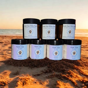 May include: A display of pet ear care products on a sandy beach. The image features several jars of "Fresh Ears" and "Fresh Ears Extra Care" ear wipes, each with a paw print logo. The jars are stacked, with the ocean and sunset in the background.