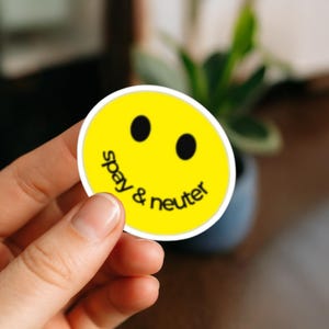 May include: A round yellow sticker with a smiling face and the words "spay & neuter" in black. The sticker has a white border and is held by a hand.