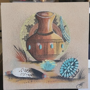 May include: A square sand art piece featuring a central brown clay pot with turquoise accents, set against a circular woven pattern. Below, a white spotted feather with a red tip and a turquoise cluster brooch rest on a textured sandy surface.