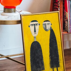 Peut inclure: Tableau encadré représentant deux personnages stylisés vêtus de noir et au visage blanc sur fond jaune. L'œuvre est dans un cadre doré et posée sur une surface en bois. Une lampe orange et un tourne-disque sont en arrière-plan.