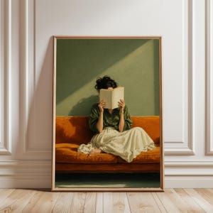 May include: A framed wall art print depicting a person with dark hair sitting on a vibrant orange sofa. The person wears a dark green long-sleeved shirt and a long cream-coloured skirt, holding an open book in front of their face against a green wall.