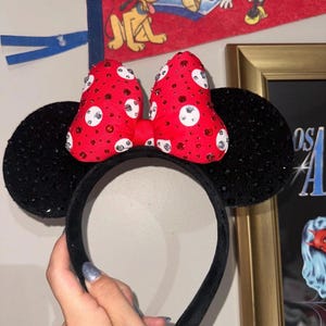 May include: Black sequined Minnie Mouse ears with a red bow adorned with white and silver rhinestones. The headband is black and velvet. A Disney-themed pennant is visible in the background.