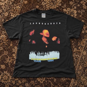 May include: A black short-sleeve t-shirt featuring the text SOUNDGARDEN at the top. The center graphic displays abstract orange and red shapes above a light blue rectangular band containing a forest silhouette and the text SUPERUNKNOWN TOUR 1994.