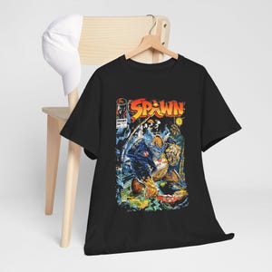 Könnte beinhalten: Schwarzes T-Shirt mit Comic-Cover-Design. Die Grafik zeigt das Wort "SPAWN" in Gelb und Orange sowie eine detaillierte Illustration einer Figur in einem blauen Kostüm. Das T-Shirt hängt an einem Holzkleiderbügel.