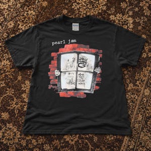 以下が含まれることがあります： 白でpearl jamと書かれた黒の半袖Tシャツ。グラフィックは赤いレンガの背景に窓枠が描かれており、両手がその枠を掴んでいる。窓のパネルの中には、黒い文字で様々な曲名が記されている。