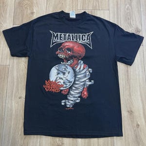 Może przedstawiać: Czarny t-shirt z nazwą zespołu "Metallica" w białej, stylizowanej czcionce. Poniżej znajduje się czerwona grafika czaszki owiniętej białymi bandażami, trzymającej globus z napisem "Wherever We May Roam".