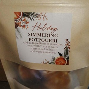 Op de afbeelding: Een beige papieren zakje met een doorzichtig venster, gevuld met potpourri om te laten sudderen. Het etiket bevat botanische illustraties, schijfjes sinaasappel en de tekst: Holiday SIMMERING POTPOURRI. Voeg alle ingrediënten toe aan een pan.