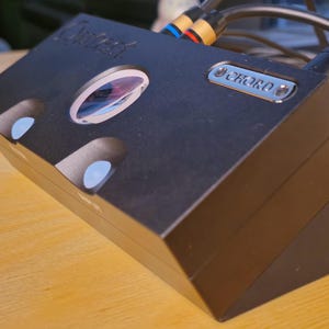 May include: Black audio component with the brand name "Chord" on a silver plate. The device has a slanted design with several circular openings and a large, oval-shaped lens. Cables are connected to the back of the device.