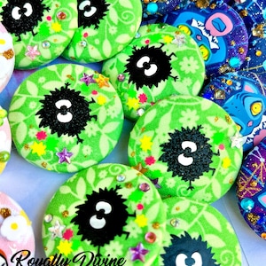 May include: A collection of round, green fabric buttons with a black, fluffy character design and the number 3 in white. The buttons are decorated with small, colorful star-shaped and gem-like embellishments. Some buttons feature a floral pattern.