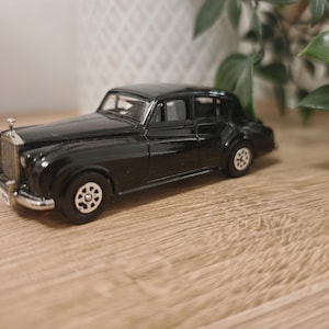 Può includere: Un'auto giocattolo nera d'epoca con dettagli argentati, tra cui la griglia e le ruote. L'auto classica ha un ornamento sul cofano ed è posizionata su una superficie di legno. L'auto giocattolo è una replica in miniatura di un veicolo di lusso.