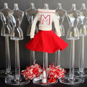 Puede incluir: Un conjunto de animadora en miniatura, que incluye un suéter blanco con una "M" roja, una falda roja, pompones, un megáfono y zapatos rojos. El conjunto se exhibe en un soporte de maniquí transparente, con otros soportes en el fondo.