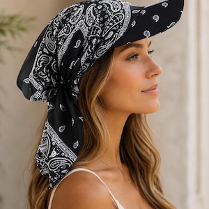 May include: A black headscarf with a white paisley pattern, featuring a structured visor brim. The fabric is tied at the back of the head, with the ends of the scarf draping down. The design combines the look of a traditional bandana with a sun visor.
