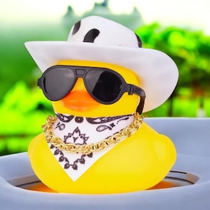 Op de afbeelding: Een gele rubberen eend met een witte cowboyhoed met zwarte vlekken, een zwarte pilotenbril, een witte bandana met een zwart paisley-patroon en een goudkleurige ketting om de nek, tegen een wazige groene achtergrond.