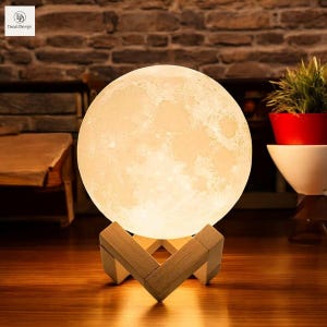 May include: A white moon-shaped lamp with a realistic moon surface texture, illuminated with a warm white light. The lamp is on a wooden stand with a geometric design.