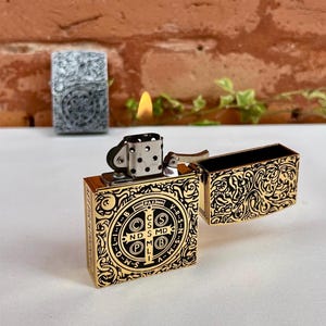 May include: Gold-colored lighter with intricate black filigree and a religious emblem. The lighter is open, revealing the flame. Another lighter with a similar design is visible in the background.