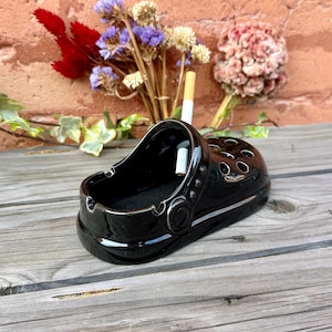 May include: Black ceramic ashtray shaped like a clog with a cigarette resting inside.