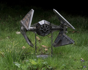 May include: A metal sculpture of a TIE fighter, a fictional spacecraft from the Star Wars franchise. The dark gray sculpture has angled solar panels and is set in a grassy area with green grass and some purple flowers.