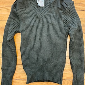 May include: A dark green V-neck sweater with ribbed knit texture. The sweater features shoulder patches and a small label near the neckline. The sleeves are long, and the garment is displayed on a wooden surface.