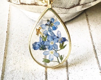 May include: A gold teardrop-shaped pendant necklace with a clear resin insert featuring pressed blue forget-me-not flowers.
