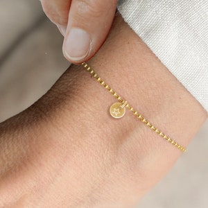 Könnte beinhalten: Ein goldenes Kettenarmband mit einem kleinen runden Anhänger. Der Anhänger hat ein Sternmuster.