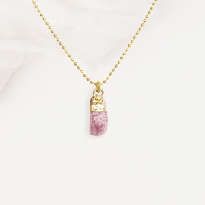 Pode incluir: Um delicado colar dourado com um pingente de turmalina rosa bruta. O pingente tem uma forma áspera e alongada, suspenso por uma corrente fina. O fundo é um tecido macio branco-sujo, criando uma estética minimalista.