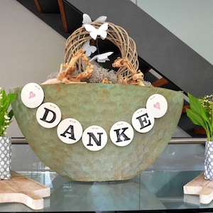 May include: A green ceramic bowl with a wreath of twigs and white butterflies, decorated with a banner that reads "DANKE" in black letters on white circles. Two white polka dot fabric containers with lily of the valley flowers are on either side of the bowl.