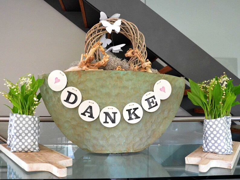 May include: A green ceramic bowl with a wreath of twigs and white butterflies, decorated with a banner that reads "DANKE" in black letters on white circles. Two white polka dot fabric containers with lily of the valley flowers are on either side of the bowl.