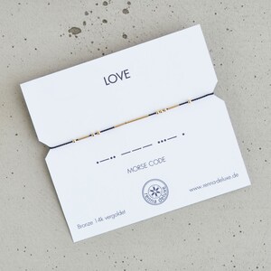 May include: A white card with the word "LOVE" printed in black. A black string with gold beads is strung across the card. The card also includes the text "MORSE CODE" and the website address "www.remarkabledeluxe.de".