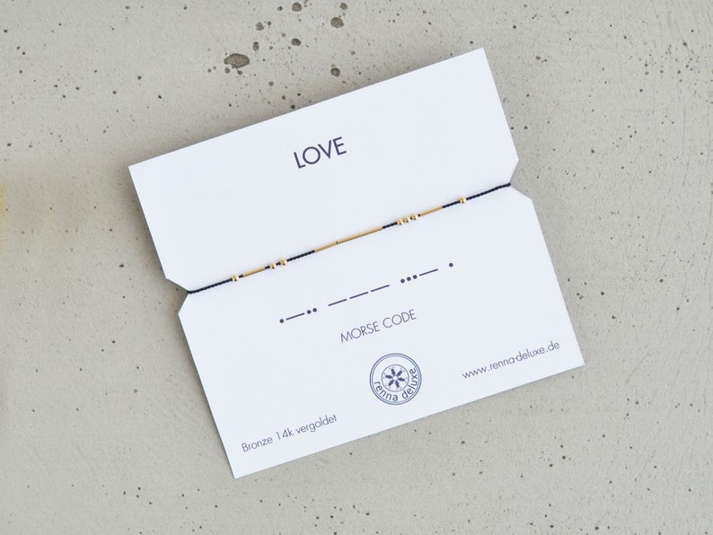 May include: A white card with the word "LOVE" printed in black. A black string with gold beads is strung across the card. The card also includes the text "MORSE CODE" and the website address "www.remarkabledeluxe.de".