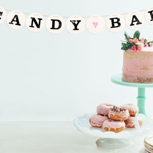 May include: A pink and white frosted cake with flowers on top sits on a mint green cake stand. A white cake stand with pink frosted donuts sits in front of the cake stand. A banner with the words "CANDY BAR" hangs above the cake stand.