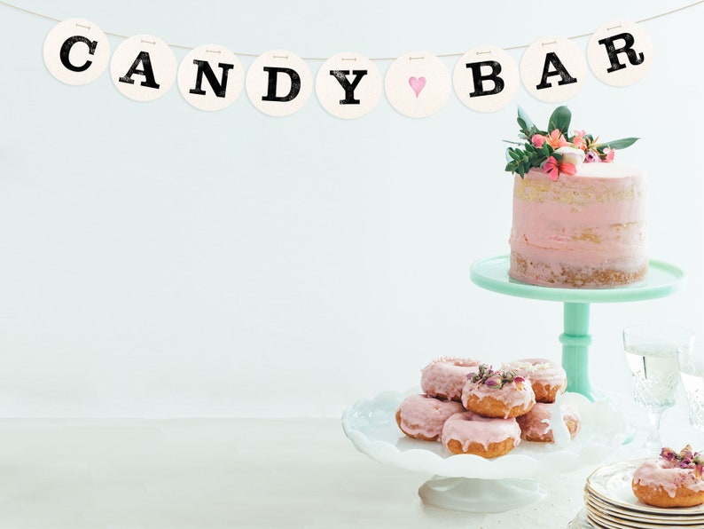 May include: A pink and white frosted cake with flowers on top sits on a mint green cake stand. A white cake stand with pink frosted donuts sits in front of the cake stand. A banner with the words "CANDY BAR" hangs above the cake stand.