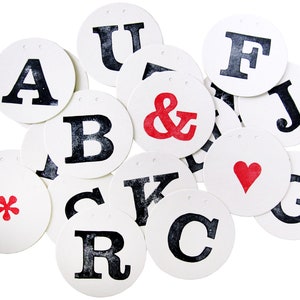 May include: A set of white circular paper tags with black letters and symbols, including A, B, C, F, G, J, K, R, U, and the ampersand symbol. One tag has a red heart, another has a red asterisk, and another has a red ampersand.