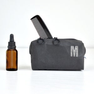 Puede incluir: Una bolsa de aseo negra con cierre de cremallera y dos anillos metálicos. La bolsa tiene una letra "M" blanca impresa en la parte delantera. Un peine negro sobresale de la parte superior de la bolsa. Una pequeña botella de vidrio marrón con un gotero está en primer plano.