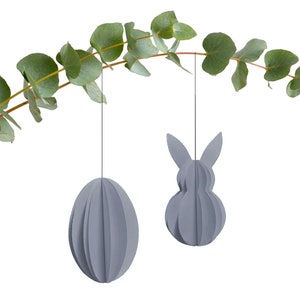 Puede incluir: Dos decoraciones de Pascua de papel gris que cuelgan de una rama de eucalipto. Una decoración es un conejo y la otra es un huevo.