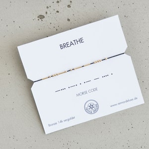 May include: A black string bracelet with gold beads spelling out "breathe" in Morse code. The bracelet is displayed on a white card with the word "breathe" and "Morse code" printed on it.