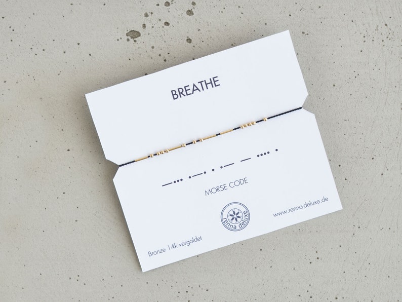 May include: A black string bracelet with gold beads spelling out "breathe" in Morse code. The bracelet is displayed on a white card with the word "breathe" and "Morse code" printed on it.