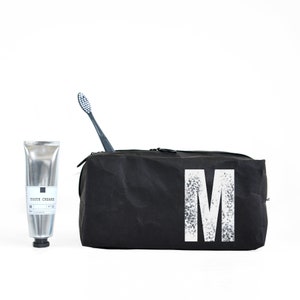 Puede incluir: Bolsa de aseo negra con una letra "M" blanca impresa en la parte delantera. Un tubo de pasta de dientes plateado y un cepillo de dientes negro están delante de la bolsa.