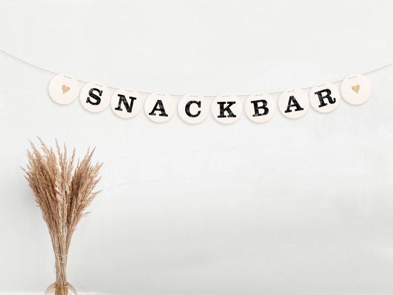 May include: A white banner with black letters spelling "SNACK BAR" and gold hearts on either end. The banner is strung on a white wall with a bouquet of dried pampas grass in a clear vase in the lower left corner.