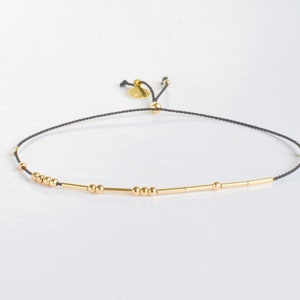 May include: A delicate black string bracelet with gold beads and bars. The bracelet is adjustable with a sliding knot.