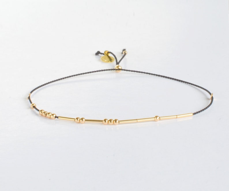 May include: A delicate black string bracelet with gold beads and bars. The bracelet is adjustable with a sliding knot.