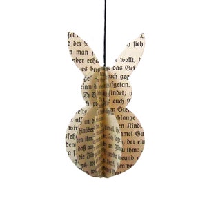 Könnte beinhalten: Ein Papier-Osterhase, hergestellt aus alten Buchseiten. Der Hase hat einen dreidimensionalen Körper und lange Ohren, die an einem schwarzen Faden hängen. Der Text aus dem Buch ist auf der Oberfläche sichtbar, was ihm ein rustikales Aussehen verleiht.