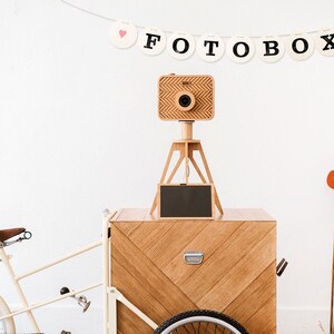 May include: A wooden photo booth on a white bicycle cart with a wooden tripod and camera. The photo booth has a black chalkboard sign and a red hat is on a wooden step ladder in the background. A banner above the photo booth reads "FOTOBOX" with pink hearts on each end.