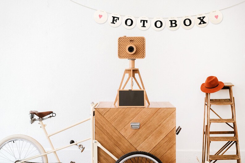May include: A wooden photo booth on a white bicycle cart with a wooden tripod and camera. The photo booth has a black chalkboard sign and a red hat is on a wooden step ladder in the background. A banner above the photo booth reads "FOTOBOX" with pink hearts on each end.